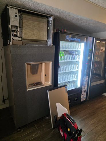Vending machines with maintenance tools and a removed panel in an indoor setting.