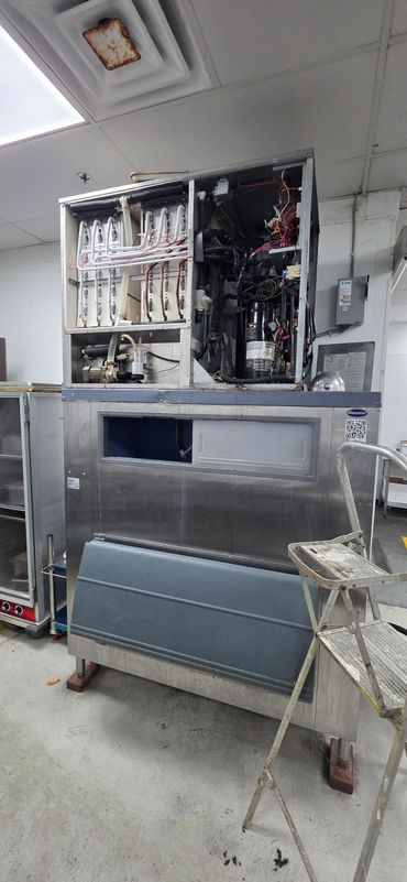 Industrial ice machine with exposed internal components and a step ladder beside it.