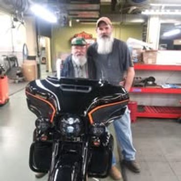 Two men standing behind a black motorcycle in a workshop.