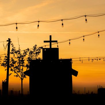 Silhouette of a church with a cross at sunset, surrounded by string lights and a tree.