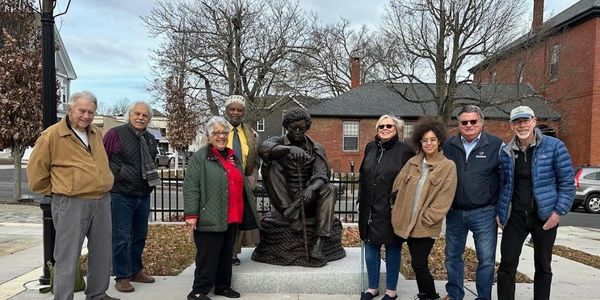 A diverse group of people posing around a statue in a park on a cloudy day.