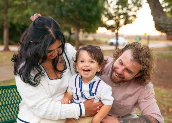 A joyful toddler smiling between two adults outdoors.