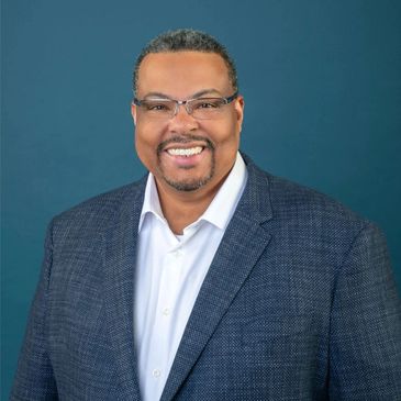 Smiling man in glasses wearing a blue blazer and white shirt against a blue background.