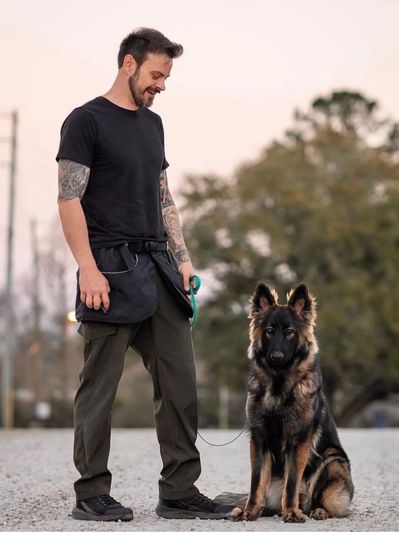 A man with a beard and tattoos stands next to a sitting German Shepherd.