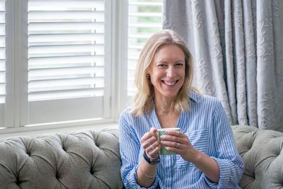 Smiling women in striped blue shirt holding mug on green sofa with white shutters and green curtains