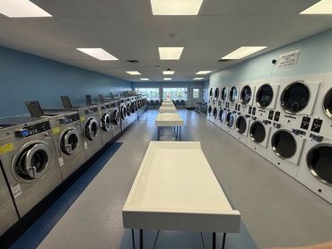 A clean, well-lit laundromat with rows of washers and dryers and folding tables.