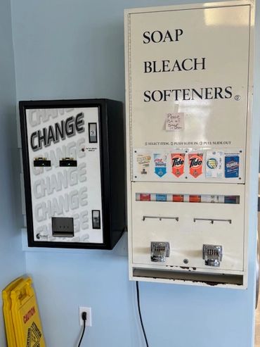 Laundry soap and change machines on a wall with a caution sign nearby.