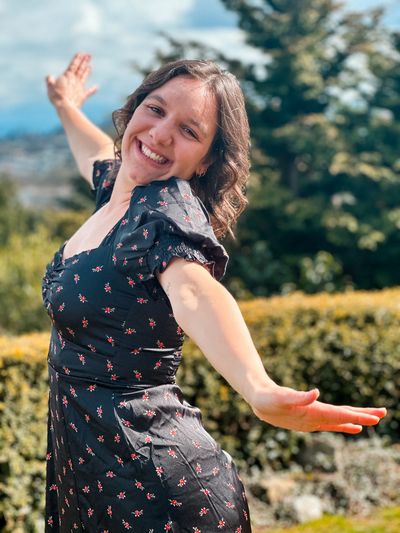 Woman joyfully posing outdoors with open arms in a floral dress.