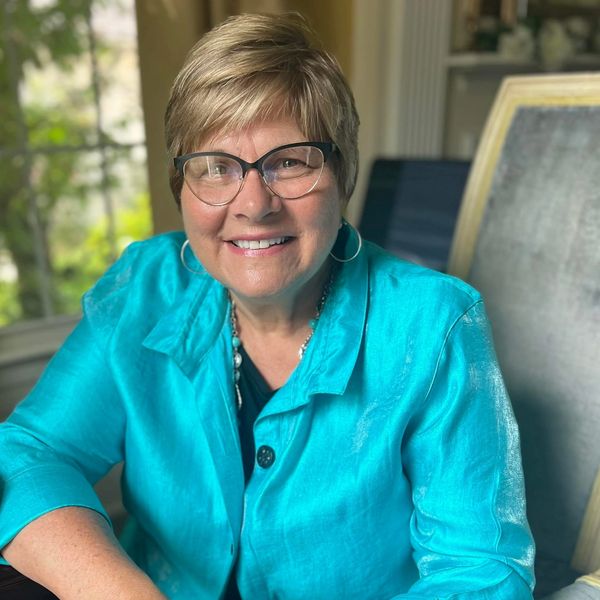Smiling woman in glasses and turquoise shirt sitting indoors.