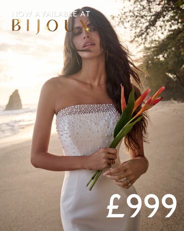 Elegant woman in pearl-adorned white dress holding tropical flowers at the beach.