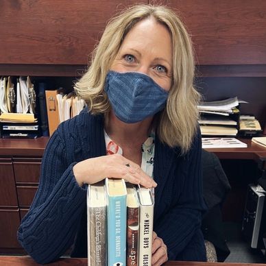 Woman wearing face mask smiling at desk while holding four books