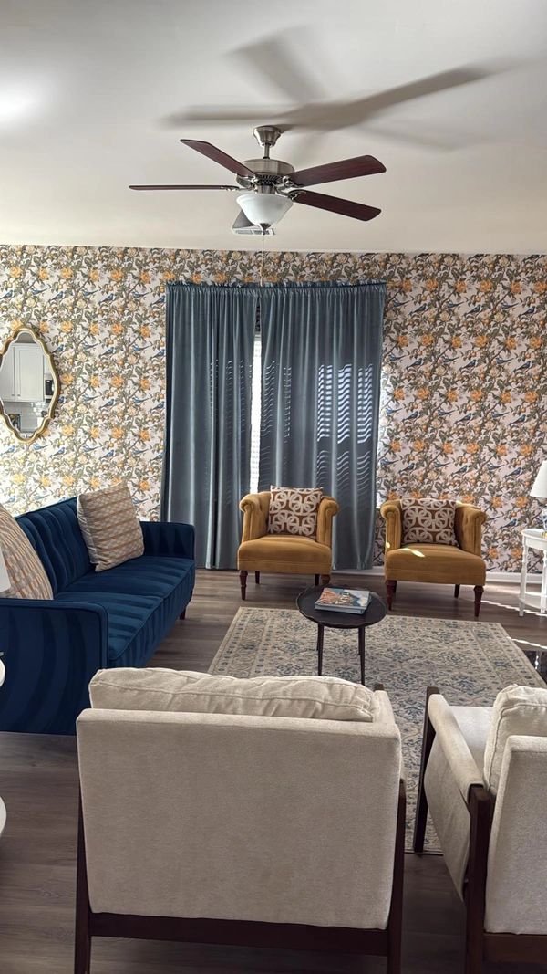 Bright living room with floral wallpaper, blue sofa, mustard chairs, and a ceiling fan.