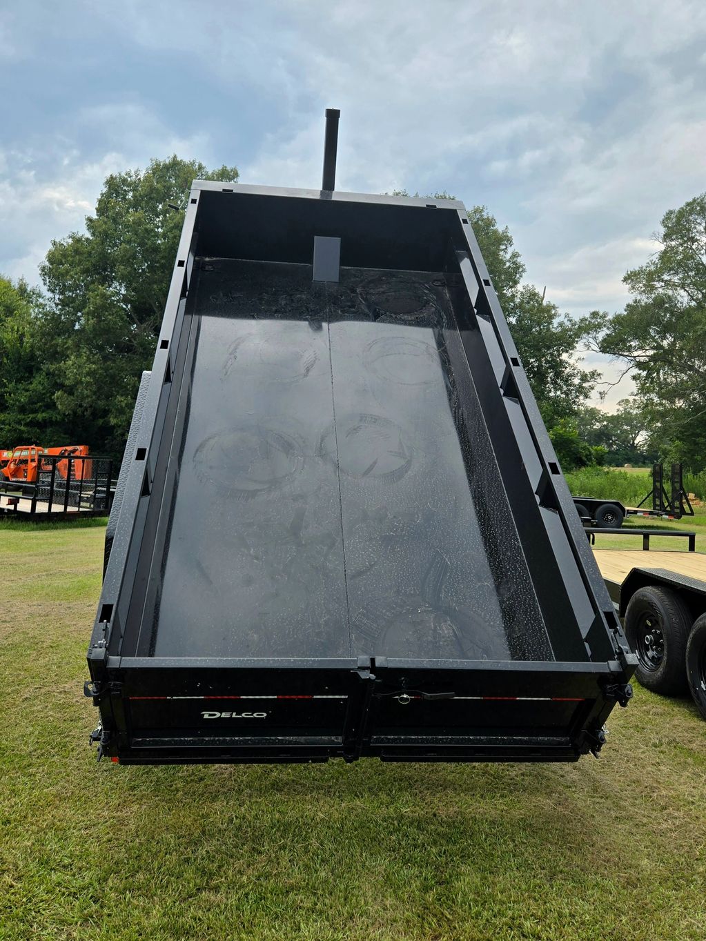 A black Delco dump trailer raised on a grassy area with trees in the background.