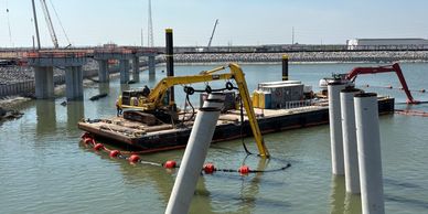 A yellow excavator on a barge working near a pier in the water.