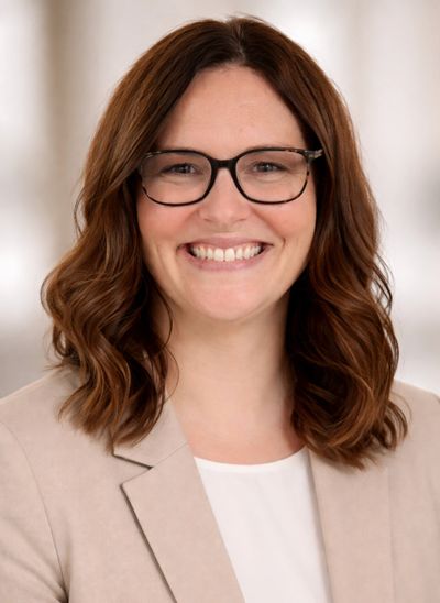 Smiling woman with glasses and wavy brown hair in a beige blazer.