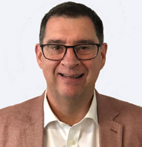 Smiling man wearing glasses and a brown blazer over a white shirt.