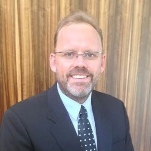 A man in a suit and tie smiling against a wooden background.