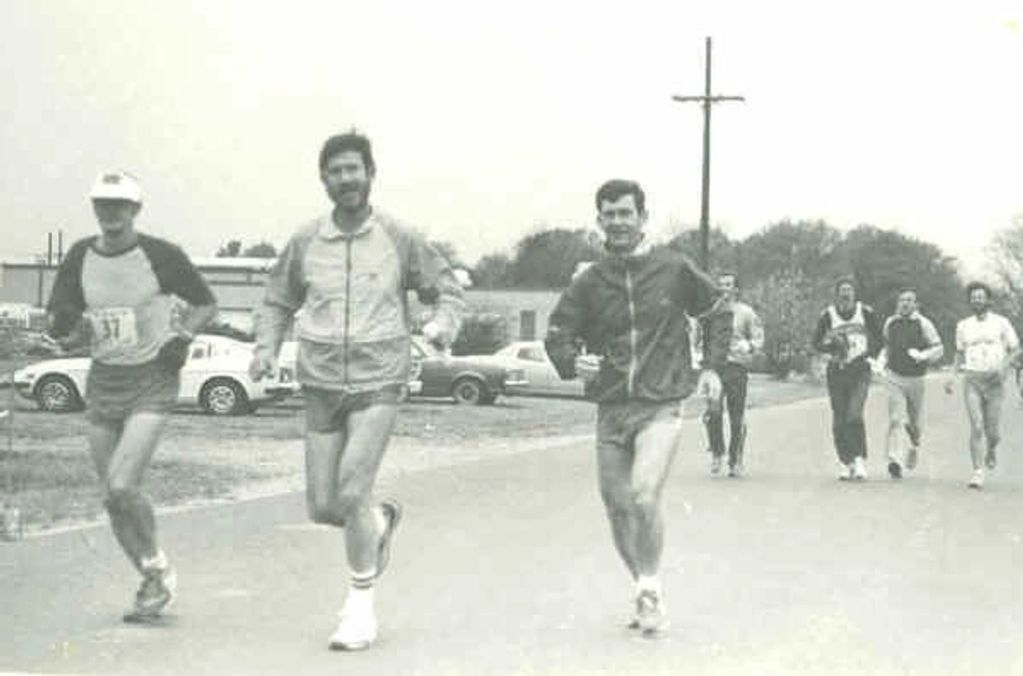 Vintage photo of runners in a road race with cars and trees in the background.