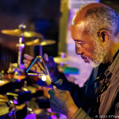 An elderly man playing a triangle in a music setting.