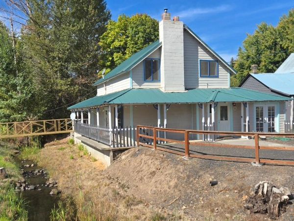 A two-story house with a green metal roof near a small creek and trees.