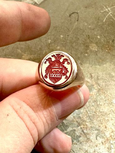 Hand holding a gold signet ring with a detailed red crest engraving.