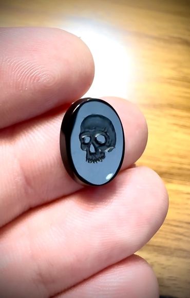 A small black oval stone with a carved skull held between fingers.