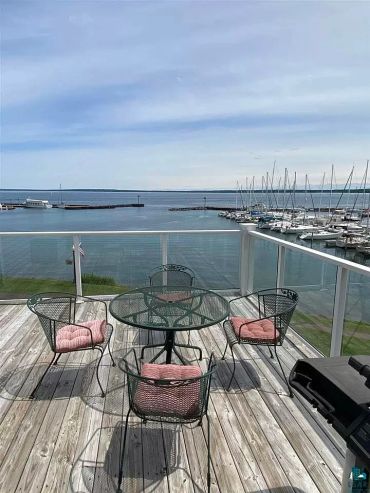 Deck with a glass table and cushioned chairs overlooking a marina and calm sea.