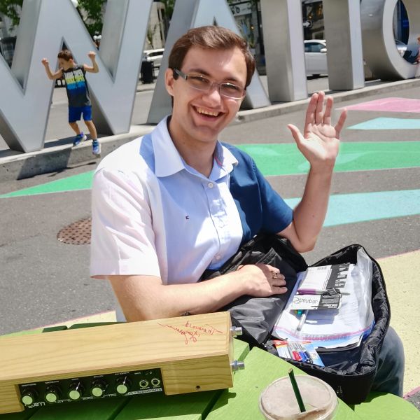 Smiling man waving while sitting at a colorful outdoor table with musical equipment and notebooks.