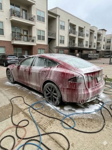 Red car covered in soap suds during wash in a residential neighborhood.