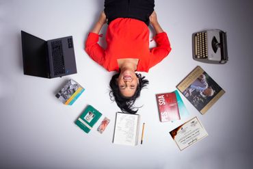 mulher deitada rodeada de livros, caderno, computador e máquina de escrever