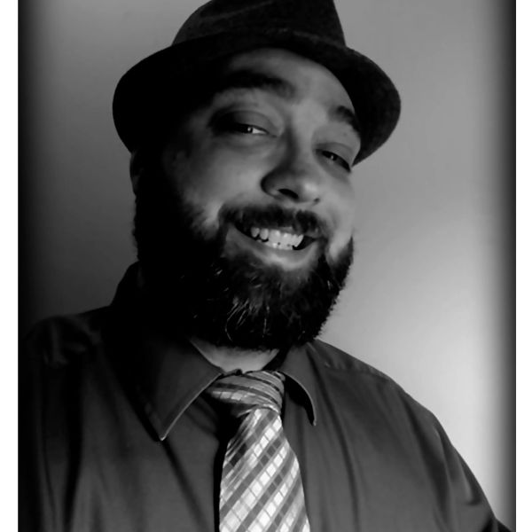 Voice talent Joshua Lute wearing a hat, dress shirt and tie smiling with a full beard.