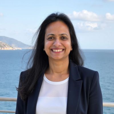 Smiling woman in blazer standing by the sea with hills in the background.