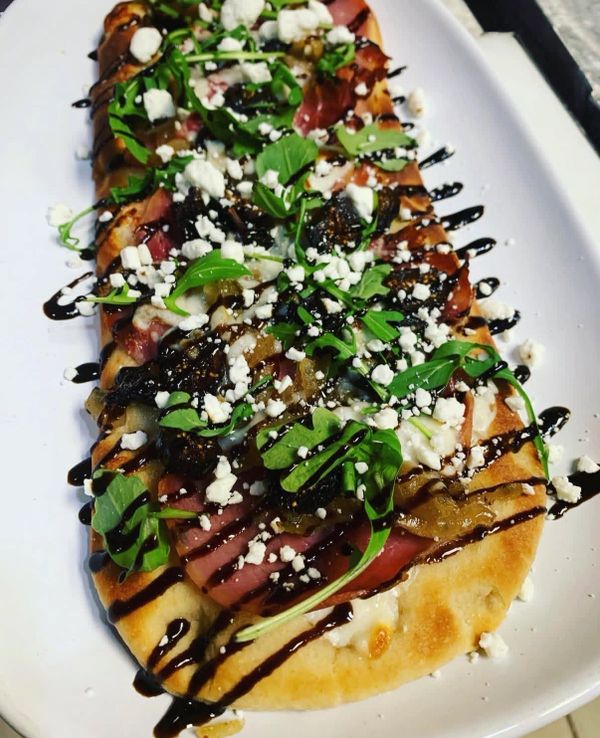 Flatbread with greens, cheese, meat, and balsamic glaze on a white plate.