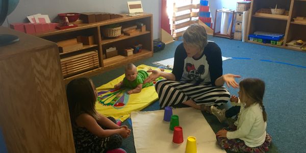 Teacher Mia playing a game with preschool students (and her baby daughter onlooking).