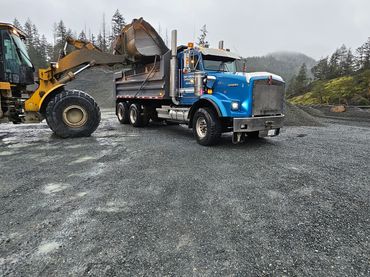 A loader fills a blue dump truck with gravel in a mountainous area.
