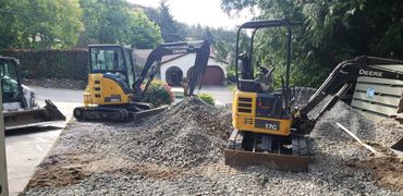 Two yellow Deere excavators working on a gravel driveway with a house in the background.