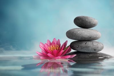 A pink lotus flower and a stack of smooth stones sitting on top of water with reflection in water.