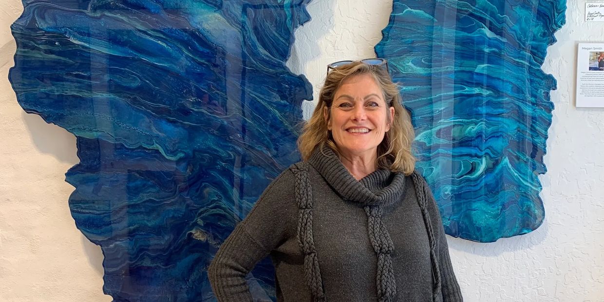 Smiling woman in a gray sweater stands in front of vibrant blue abstract wall art.