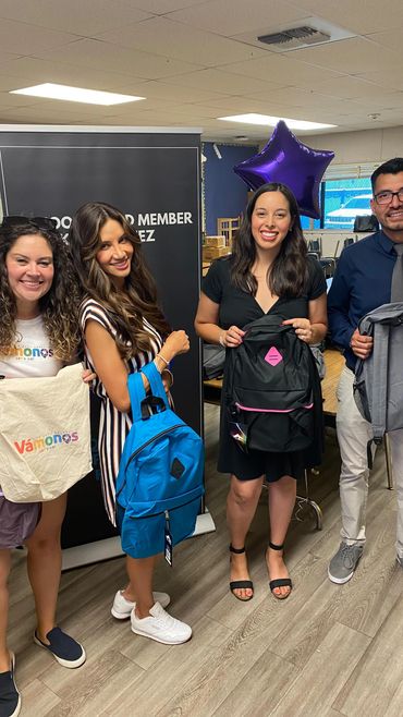 Four smiling adults holding backpacks in a classroom setting with a purple star balloon.