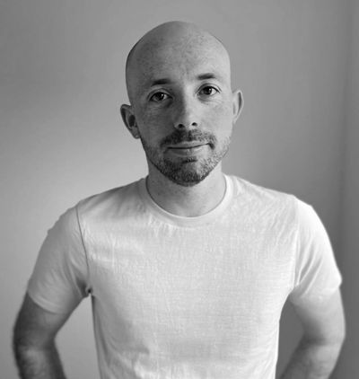A bald man with a trimmed beard wearing a light gray t-shirt, standing against a plain background.