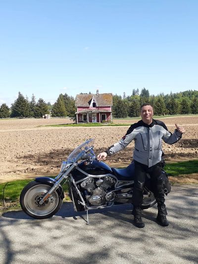 Man in motorcycle gear posing with bike near a rural house and field.