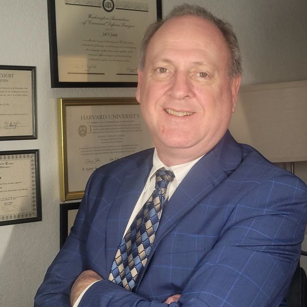 Man smiling in a blue checkered suit with framed certificates behind him.