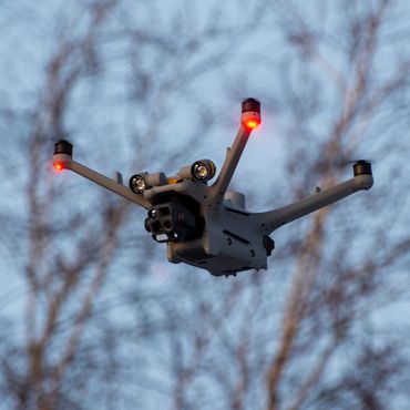 A drone with red lights flying outdoors against a backdrop of trees and sky.
