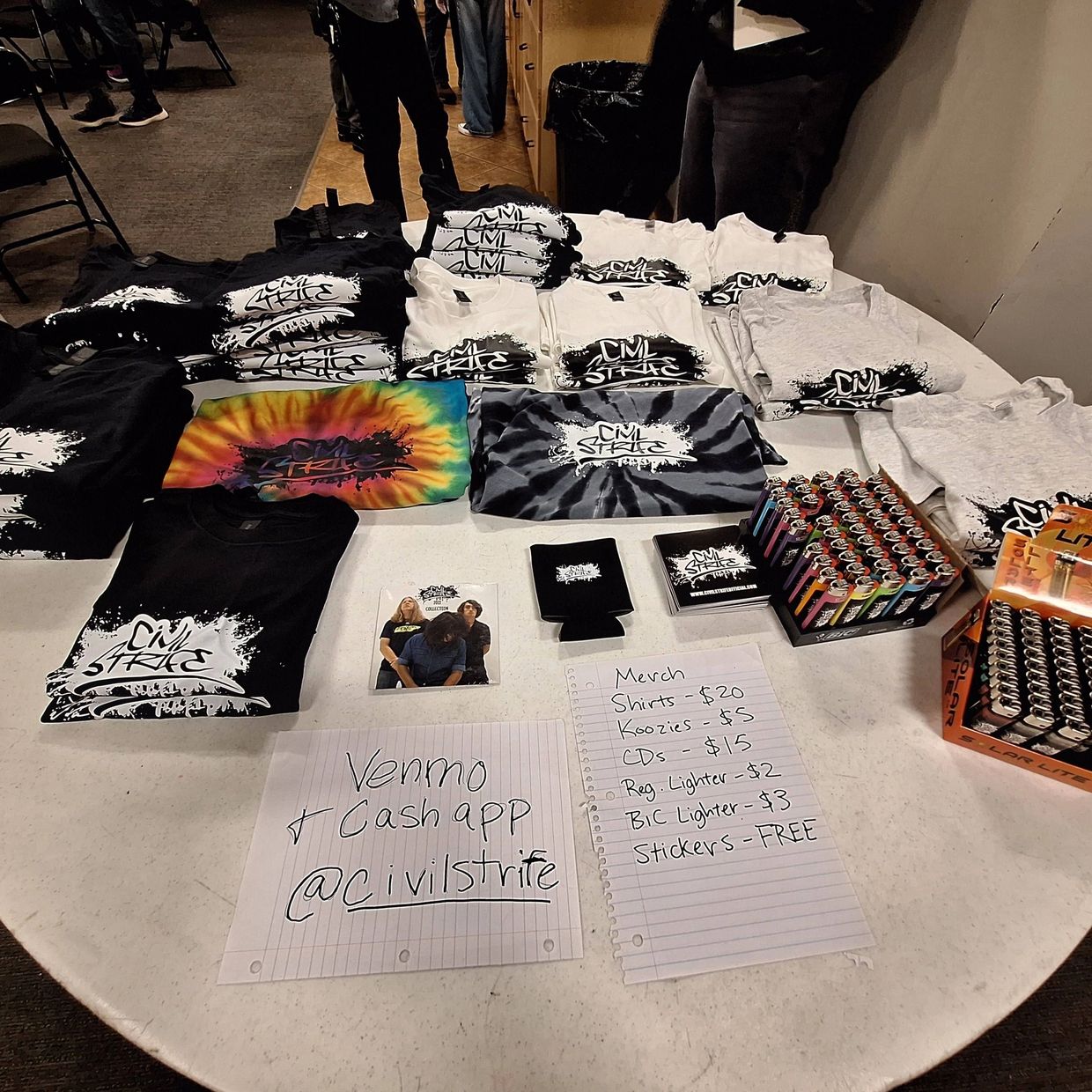 Merchandise table with shirts, CDs, lighters, and handwritten price list for Civil Strife band.