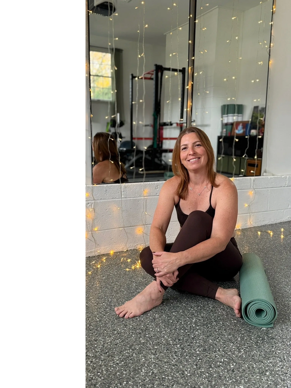 Woman in workout clothes sitting on the floor next to a yoga mat with fairy lights. Personal Training for women. Nutrition coaching for women. Private yoga instruction for women. Physical therapy background.