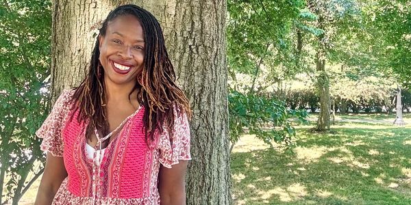 Woman in a pink patterned dress smiling by a large tree in a sunny park.