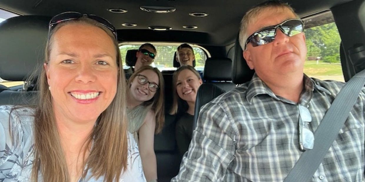 A happy family of six smiling inside a car on a sunny day.