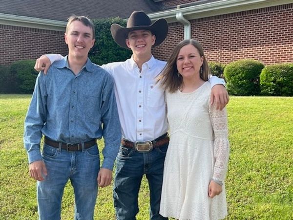Three smiling young adults standing outside in casual and western attire.
