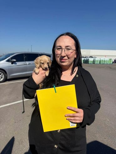 Woman holding a small puppy and a yellow folder in a sunny parking lot.