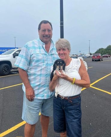 A smiling couple holding a small dog in a parking lot.